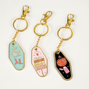 Pode incluir: Tr&ecirc;s chaveiros de motel dourados com designs diferentes. O primeiro chaveiro tem um fundo turquesa com o texto "Kickin' Boots" e um par de botas de cowboy rosa. O segundo chaveiro tem um fundo rosa com o texto "Check-in Enemies Check-out Lovers". O terceiro chaveiro tem um fundo preto com o texto "Will they want tips?" e um isqueiro rosa com chamas.