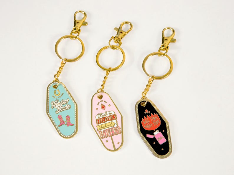 Pode incluir: Tr&ecirc;s chaveiros de motel dourados com designs diferentes. O primeiro chaveiro tem um fundo turquesa com o texto "Kickin' Boots" e um par de botas de cowboy rosa. O segundo chaveiro tem um fundo rosa com o texto "Check-in Enemies Check-out Lovers". O terceiro chaveiro tem um fundo preto com o texto "Will they want tips?" e um isqueiro rosa com chamas.