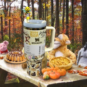 Könnte beinhalten: Ein Edelstahl-Stanley-Becher mit Winnie-the-Pooh-Figuren. Der Becher hat einen cremefarbenen Griff und Deckel sowie einen bienenförmigen Strohhalm-Aufsatz. Das Design zeigt Illustrationen von Puuh und seinen Freunden, mit dem Text "Winnie the Pooh".