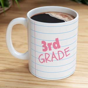 May include: White ceramic coffee mug with a blue and white lined design. The mug has a pink inscription that reads "3rd GRADE".