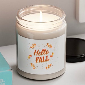 Puede incluir: Una vela encendida en un tarro de vidrio transparente con tapa negra. La etiqueta dice "Hello Fall" en naranja y amarillo, rodeada de ilustraciones de hojas de otoño. La vela está sobre una superficie blanca.