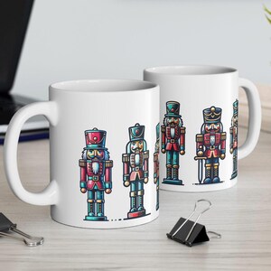 Może przedstawiać: Biały ceramiczny kubek z kolorową ilustracją żołnierza dziadka do orzechów. Projekt zawiera czerwone, niebieskie i złote akcenty. Kubek ma duży uchwyt i nadaje się do gorących lub zimnych napojów.
