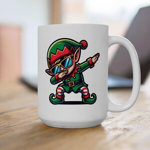 May include: White ceramic mug with a cartoon elf in a green and red outfit, sunglasses, and a dab pose. The elf has a green hat with a red trim and a red pom-pom. The mug has a white handle and is on a wooden surface.