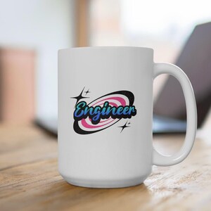 May include: White ceramic coffee mug with a pink and blue retro design featuring the word "Engineer" in a stylized font. The design includes a black oval with a pink and white swirl inside and two small stars.