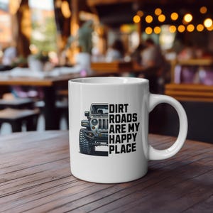 Könnte beinhalten: Weiße Keramiktasse mit einer schwarz-grauen Jeep-Illustration und dem Text "Dirt Roads Are My Happy Place". Die Tasse hat einen gebogenen Henkel. Sie steht auf einer Holzoberfläche.