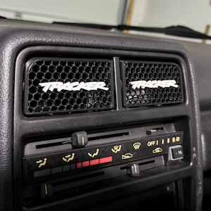 May include: A close-up of a black car dashboard showing two air vents with a honeycomb design. The word "TRACKER" is displayed in white across each vent. Below the vents is the car's climate control panel.