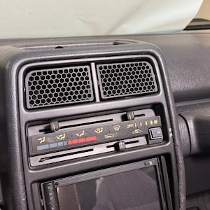 May include: Close-up of a car dashboard with air vents and climate control buttons. The vents have a honeycomb pattern. The control panel features sliders and icons for fan speed, temperature, and air direction. A car stereo is visible below.