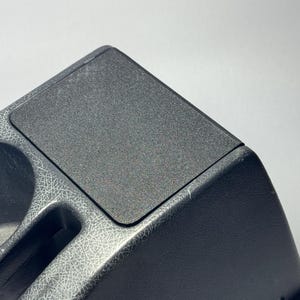 Pode incluir: Console de carro preta com uma almofada retangular texturizada e dois porta-copos. A consola tem um design rachado nas laterais e um acabamento liso e fosco na parte superior.