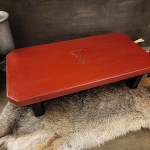 May include: A rectangular, red-painted wooden altar table with black legs. The tabletop features a carved symbol. The table is displayed on a fur-like surface, with a metal chalice and pine cone in the background. The altar table is a decorative item.