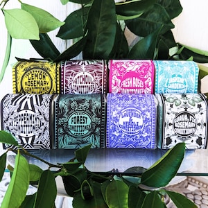 May include: Eight colorful bars of solid shampoo with different scents and names. The bars are arranged in a row and feature a variety of designs and patterns. The scents include Lemon Rosemary, Patchouli, Fresh Roses, Jasmine Gardenia, Peppermint, Forest, Lavender & Neroli, and Guava, Fig & Rosemary.