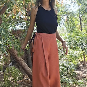 May include: A black sleeveless top paired with high-waisted, wide-leg trousers in a burnt orange hue. The trousers feature a wrap-style front, a side tie closure, and a contrasting dark vertical stripe along the side seam, creating a clean silhouette.