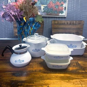 Puede incluir: Colección de utensilios de cocina CorningWare blancos vintage con un estampado floral azul. Incluye una tetera, fuentes para horno con tapas y fuentes rectangulares. El conjunto está dispuesto sobre una superficie de madera.