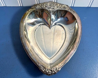 May include: Silver-toned heart-shaped dish with a floral design on the rim. The dish has a heart-shaped indentation in the center and a decorative edge. The dish is made of metal and has a reflective surface.