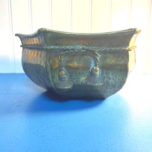 May include: A brass planter with a rope design around the top and tassels on the sides. The planter has a distressed finish.