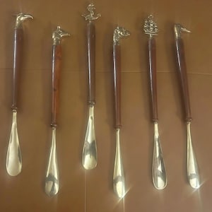 May include: A collection of six shoehorns with brass-colored blades and dark wooden handles. The handles are topped with decorative brass animal and nautical-themed designs, including a bird, horse, anchor, dog, ship, and another bird.