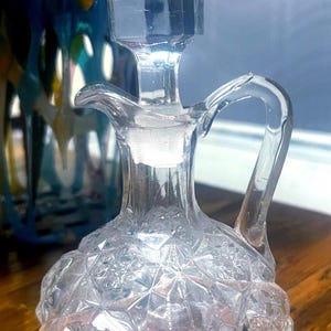 Puede incluir: Aceitera de cristal transparente con tapón y asa facetados. La aceitera tiene un patrón de diamantes en la base y un asa curvada. El fondo incluye un objeto de vidrio azul y multicolor borroso. La aceitera está sobre una superficie de madera.