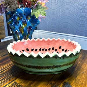 May include: A ceramic bowl designed to resemble a watermelon, featuring a pink interior with black seed details. The exterior is green with dark brown stripes, and the rim has a zig-zag pattern. A blue vase with flowers is in the background.