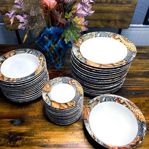 Op de afbeelding: Stapels witte keramische borden en kommen met een safari-dierrandontwerp. De borden en kommen zijn gerangschikt op een donkere houten tafel, met een kleurrijk bloemstuk op de achtergrond.