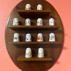 Könnte beinhalten: Ein ovales Holzregal mit mehreren kleinen Regalen, das eine Sammlung dekorativer Fingerhüte präsentiert. Die Fingerhüte sind weiß mit floralen Designs und goldenen Verzierungen. Das Regal ist an einer rosa Wand befestigt.