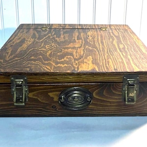 Op de afbeelding: Een houten kist met een scharnierend deksel en messing beslag. De kist heeft een donkerbruine vlek en een nerfpatroon. De kist heeft twee messing sloten en een messing handvat.