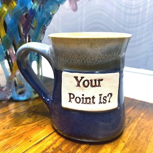 Peut inclure: Mug en céramique avec une base bleu foncé et un bord beige. Le mug présente une étiquette rectangulaire avec le texte "Your Point Is ?" en gras. La tasse a une anse incurvée.