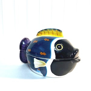 Peut inclure: Un plat de service en céramique en forme de poisson avec un motif bleu, jaune, blanc et noir. Le plat a un couvercle et est parfait pour servir des en-cas ou des amuse-gueules.