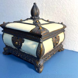 May include: A white and gold decorative box with a square shape and a pointed lid. The box has intricate details and a gold trim.