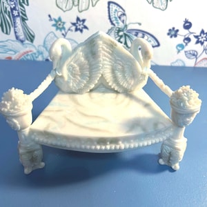 May include: A white decorative swan-shaped figurine with a bed-like structure. The figurine features two swans with outstretched wings, holding up a platform with floral accents on each side. The base is a light blue color.