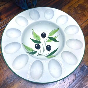 May include: White ceramic serving platter with a green rim and 12 oval compartments. The center features a hand-painted olive branch design with black olives and green leaves. A decorative piece for serving appetizers.
