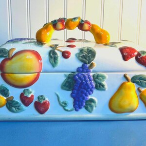 May include: A white ceramic bread box with a fruit design. The box has a handle shaped like a bunch of grapes. The fruit design includes pears, apples, grapes, strawberries, and cherries.