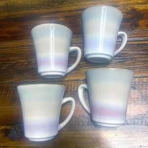 May include: Four ceramic mugs with a gradient color scheme. The mugs transition from a light blue at the top, through a beige middle, to a lavender base. Each mug has a curved handle and a slightly flared shape.
