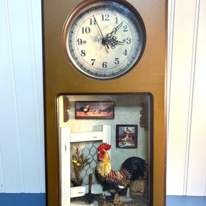 May include: A rectangular wooden clock with a round clock face at the top. The clock face has black hands and Roman numerals. Below the clock face is a diorama scene featuring a rooster, a white fence, and small paintings.