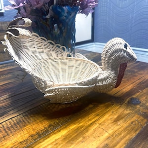 May include: A white wicker turkey-shaped basket with a red and white stripe on the neck. The basket is sitting on a wooden surface.