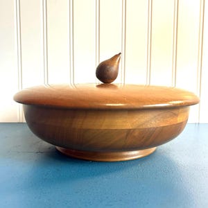 May include: A wooden bowl with a lid and a small pear-shaped knob on top. The bowl is made of light brown wood and has a smooth finish.