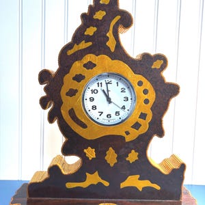 May include: A brown and gold wooden clock with a unique, abstract design. The clock face is white with black numbers and hands.