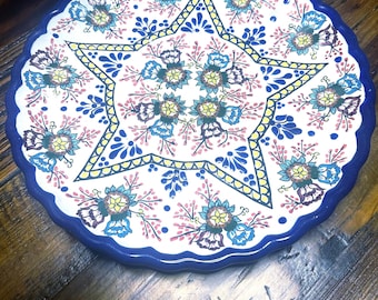 May include: A round ceramic plate with a scalloped edge. The plate has a star design with floral patterns in blue, yellow, and pink on a white background. The rim is a solid blue. The plate is likely 25-30 cm in diameter.