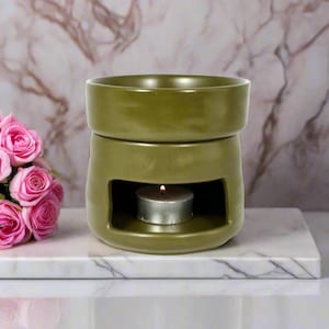 May include: A green ceramic wax melt warmer with a small tea light candle in the centre. The warmer is sitting on a white marble surface.
