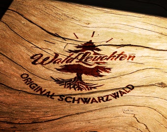 Puede incluir: Un diseño quemado en madera sobre una superficie de madera. El diseño presenta un árbol estilizado con el texto "Wald Leuchten Original Schwarzwald".