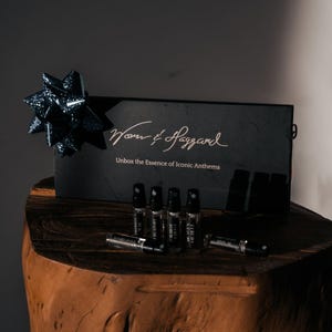 May include: A black box with the text "Wom & Haggard" and "Unbox the Essence of Iconic Anthems" on it. The box is sitting on a wooden surface with a blue bow on top. There are several small glass bottles with black caps on the surface.