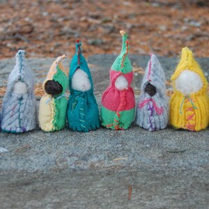 May include: A row of ten knitted dolls, each wearing a different colored hooded garment. The dolls are arranged in a line on a gray stone surface.