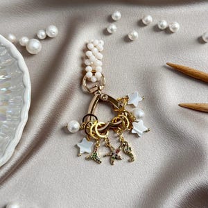Op de afbeelding: Een goudkleurige sleutelhanger met een cluster bedels, waaronder witte stervormige onderdelen, een zeester, een zeepaardje en een vissenstaart, allemaal geaccentueerd met kleine gekleurde steentjes. Aan de bovenste sluiting zit een streng witte kralen.