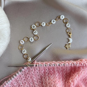 以下が含まれることがあります： 1から9まで番号が付けられた白いビーズが付いた金色のチェーンで、月と星のチャームが付いています。チェーンは、銀色の編み針が付いたピンク色の編まれた生地に付けられています。