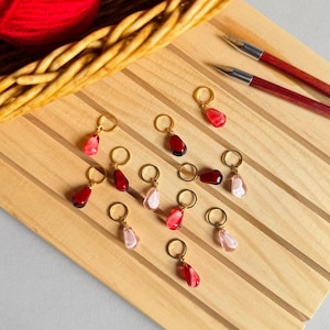 Puede incluir: Marcadores de punto rojos y rosas en forma de lágrima con anillos dorados, dispuestos sobre una superficie de madera. También hay una cesta con lana roja y dos agujas de tejer de madera. Diseñados para proyectos de punto y ganchillo.