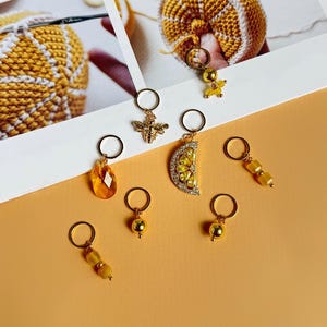 Könnte beinhalten: Ein Set aus acht gelben und goldenen Maschenmarkierern mit verschiedenen Anhängern, darunter eine Biene, eine Zitronenscheibe und Perlen. Die Marker sind auf einer hellorangen Oberfläche angeordnet.