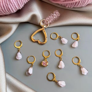 Op de afbeelding: Een verzameling goudkleurige steekmarkeerders en een hartvormige sluiting met een roze kralenbedel. De steekmarkeerders hebben een gouden ring met een druppelvormige, wit-roze bedel. Roze garen staat op de achtergrond.