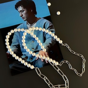 May include: A necklace featuring a combination of white round pearls and silver-toned metal paperclip links. The jewellery piece is displayed over a photograph of a person wearing a blue cardigan and a pearl necklace, with loose pearls nearby.
