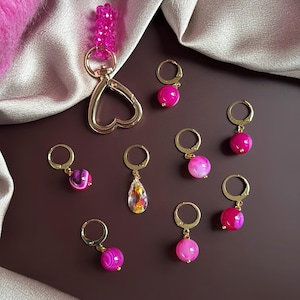 May include: A collection of gold-toned earrings and a keychain with pink gemstone beads. The earrings feature various pink and clear gemstone drops and round beads. The keychain has a heart-shaped clasp and a pink beaded accent.