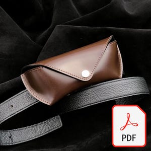 May include: A smooth brown leather glasses case with a triangular flap and a silver metal snap button closure, positioned resting on top of a black textured leather belt with visible stitching and a series of adjustment holes along the strap.