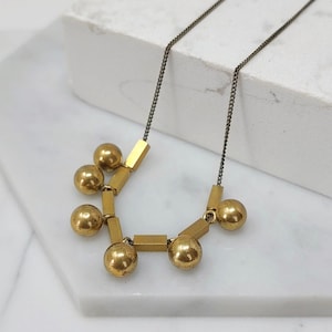 Puede incluir: Un collar de oro con un diseño geométrico que presenta formas redondas y rectangulares. El collar está hecho de latón y tiene una cadena negra.