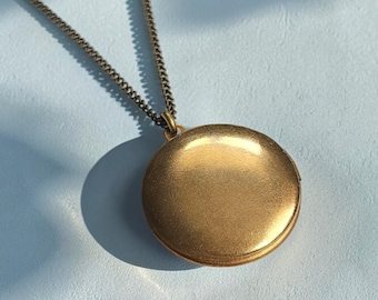 May include: A round, matte gold-toned locket pendant suspended from a matching thin metal chain. The locket features a smooth, plain circular surface with a subtle metallic sheen, reflecting light across its face while hanging from a delicate link chain.